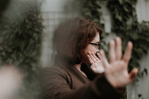 Женщина в очках стоит, отвернувшись от камеры и закрывая лицо руками